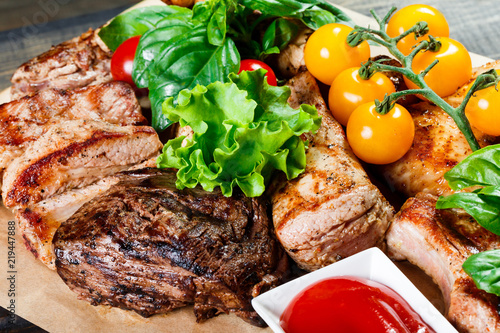 Assorted delicious grilled meat and vegetables with fresh salad and bbq sauce on cutting board on wooden background close up