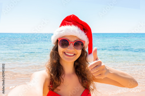 young girl in santa hat shows thumb upand having fun taking smartphone selfie pictures of herself. travel winter holidays photo