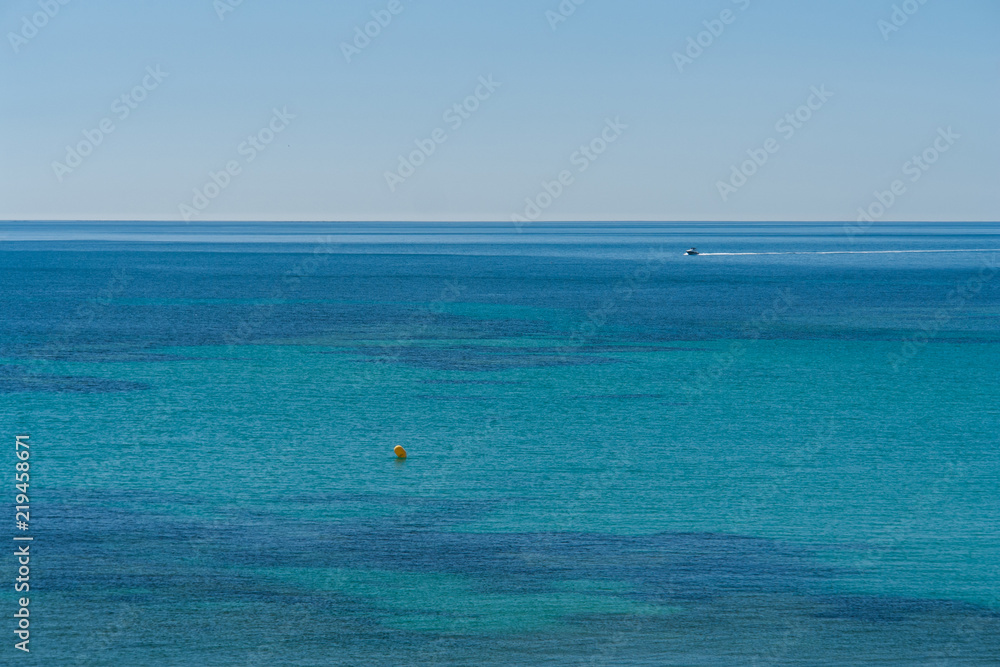 Different blue shades at a beach of the Mediterranean Sea with a yacht at the background on a summer day with clear sky