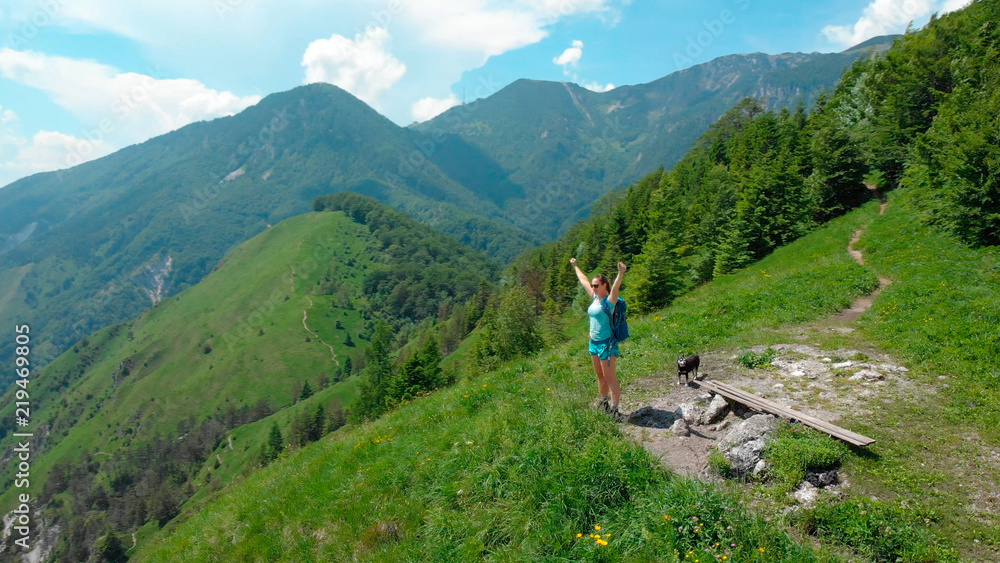 Fototapeta premium AERIAL: Joyful female hiker celebrates a successful ascent of mountain in Alps.
