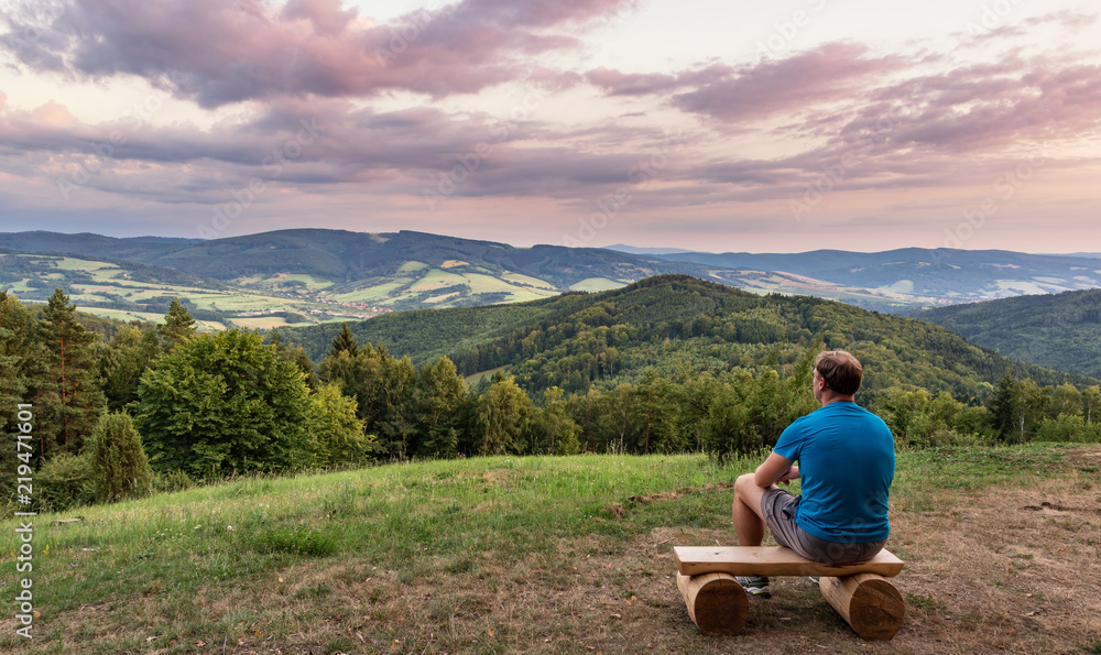 A man is looking at a landscape of mountains, hills, meadow, forest during a cloudy sunset. Calm and peaceful looking at the beauty of a country.