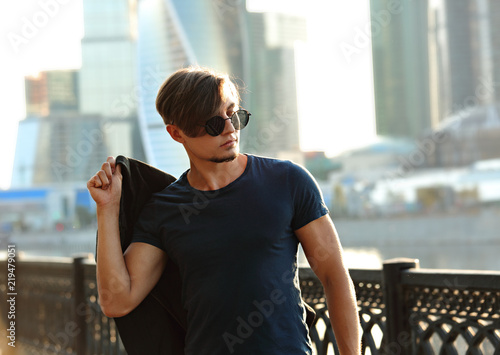 Handsome young man posing in fashion sun glasses and holding the jacket behind hisself and looking on city center background photo