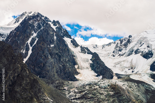 Snowy mountain top under blue cloudy sky. Water flows from icy bright rocks. Dark steep mountainside. Atmospheric landscape of majestic nature.