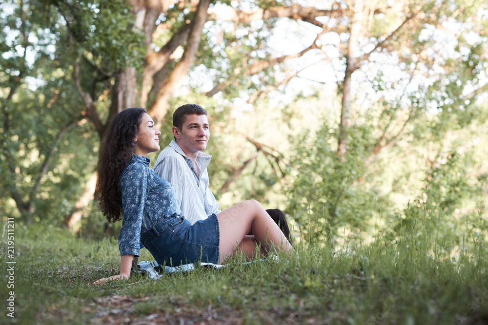 young couple sitting on the grass in the forest and looking on sunset, summer nature, bright sunlight, shadows and green leaves, romantic feelings