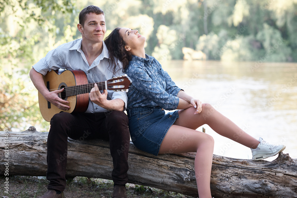 young couple sitting on a log by the river and playing guitar, summer nature, bright sunlight, shadows and green leaves, romantic feelings