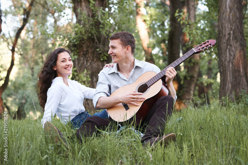 young couple sitting in the forest and playing guitar, summer nature, bright sunlight, shadows and green leaves, romantic feelings