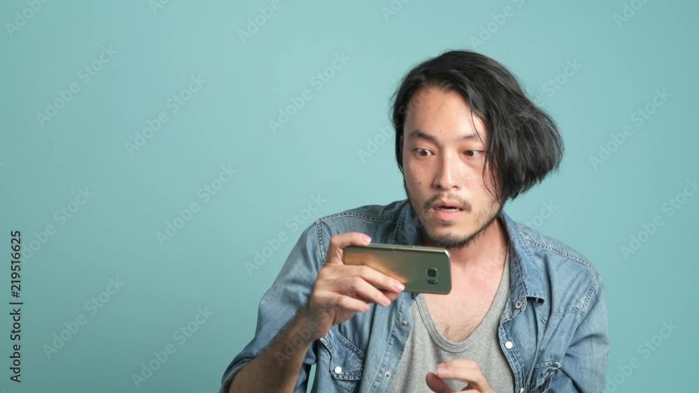 Young bearded male hipster looking at his phone to find great new from mobile message application. Bored Asian man looking at mobile phone and extremely happy as he see good news with blue background. Young bearded male hipster looking at his phone to find great new from mobile message application. Bored Asian man looking at mobile phone and extremely happy as he see good news with blue background.