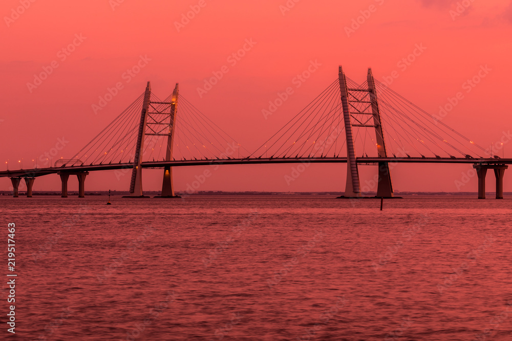 Obraz premium The bridge of circle highway road over Neva river near the mouth of it in the gold hour during the sunset. Evening view on the buildings of Petersburg city and the Finish gulf