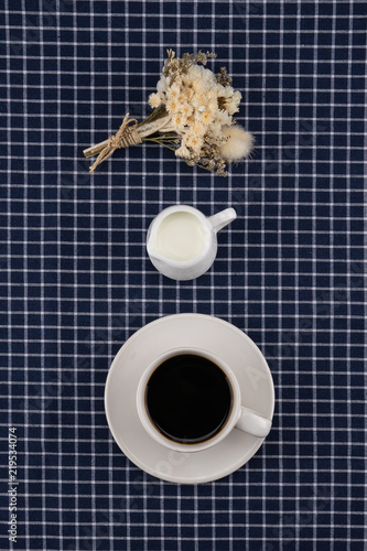 A cup of coffee, milk and white flower bouquet on dark blue table cloth