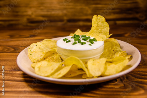 Ceramic plate with potato chips and glass bowl with sour cream on wooden table