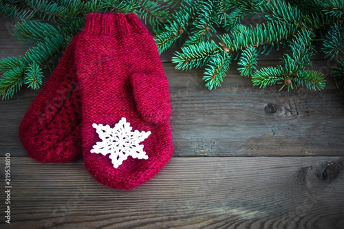 Red wool mittens with white cotton crocheted snowflake near green christmas fur tree on the rustic wooden background. Toned. Christmas card, postcard concept.