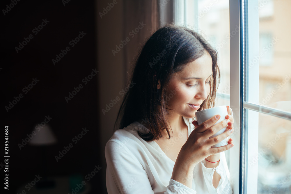 Happy beautiful woman drinking hot coffee looking on window at home