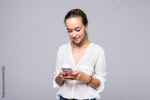 Attractive smiling girl standing against gray background and typing sms on her smartphone