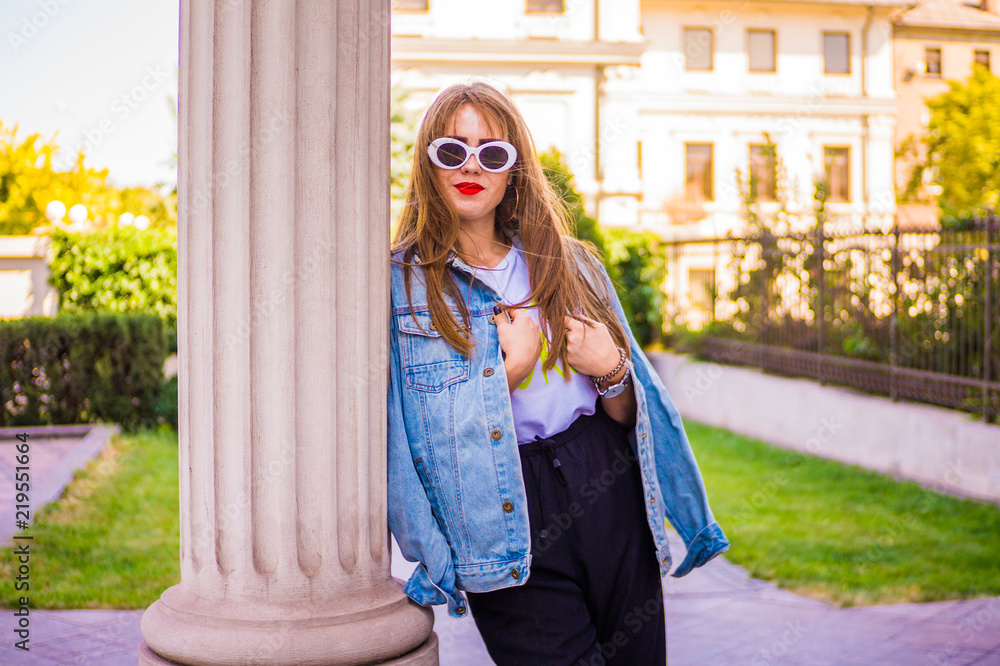 Fototapeta premium Fashionable girl in broad trousers and a white T-shirt in sneakers and a denim jacket, and fashionable glasses with a wide white frame, stylishly dressed, poses and shows her outfit
