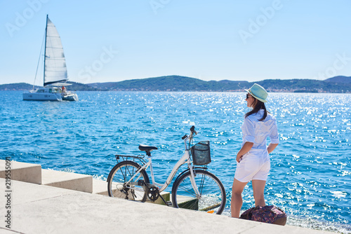 Back view of slim tourist woman in white clothing and sunglasses resting at bicycle and backpack on paved stone sidewalk stairs on clear blue sea water, sailing cruse yacht and mountains background.
