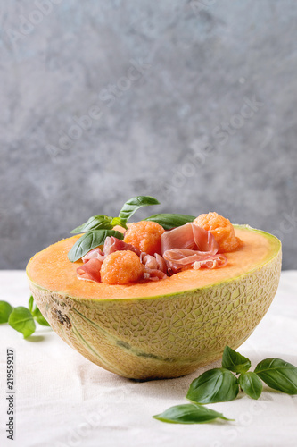 Melon and ham or prosciutto salad served in half of Cantaloupe melon, decorated by fresh basil standing on white tablecloth.