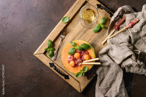 Melon and ham salad served in half of Cantaloupe melon with fresh basil and grissini bread on wooden serving tray over dark brown texture background with glass of white wine. Flat lay, space