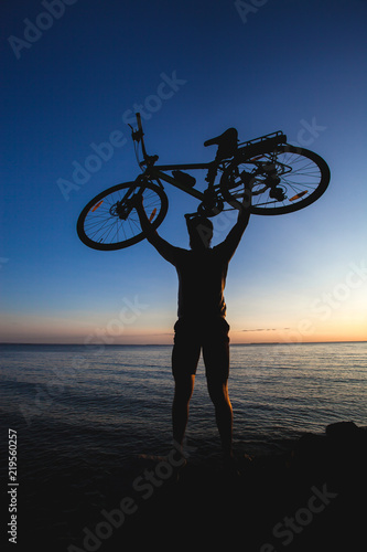 silhouette of a cyclist is holding his bicycle over himself on the sunset background