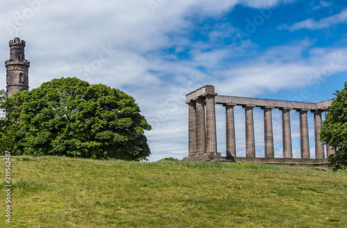 Edinburgh, Scotland, UK - June 13, 2012: Nelson and National Monuments on Calton Hill captured between green trees behind green lawn and under blue cloudy sky.