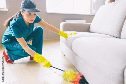 Cleaner is sitting in squad position and holding dust brush. She is cleaning underneath sofa. Girl is serious and conentrated.