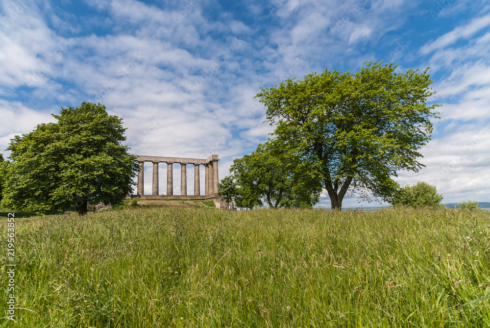 Edinburgh, Scotland, UK - June 13, 2012: National Monument is series of brown pilars against blue sky with white clouds. Green grass up front. Two trees. Edinburgh, Scotland, UK - June 13, 2012: National Monument is series of brown pilars against blue sky with white clouds. Green grass up front. Two trees.