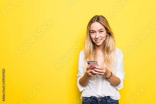 Portrait of young girl holding mobile phone while standing isolated over yellow background