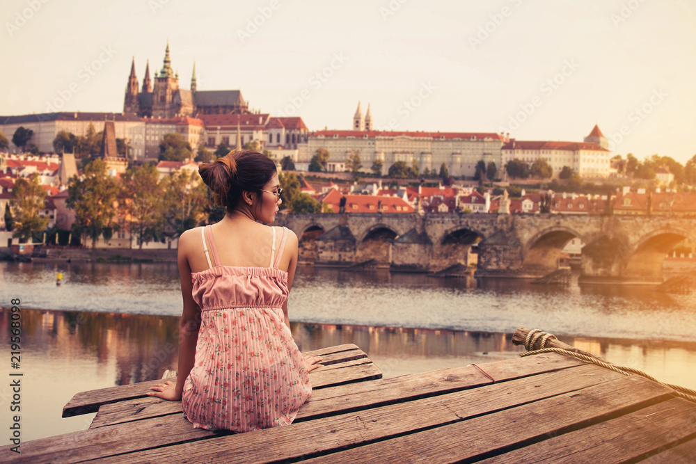 Young woman sitting on wood pier looking Prague Castle, Prague river Vltava and many famous Prague sights and historic architecture, back view. - obrazy, fototapety, plakaty 