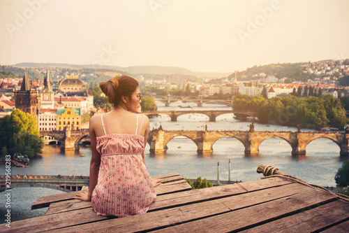 Obraz na plátně Young woman sitting on wood pier looking Prague Castle, Prague river Vltava and many famous Prague sights and historic architecture, back view