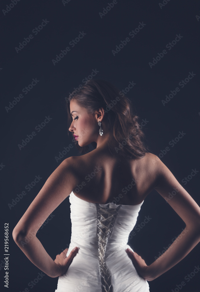 Beautiful slender young woman in white corset wedding or evening dress, stands with her back on a dark background, beauty and fashion, glamor