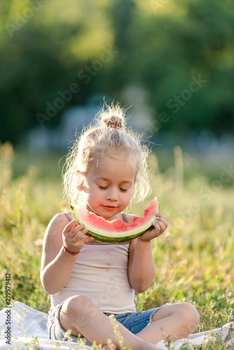 Little blond girl eating watermelon in the park.