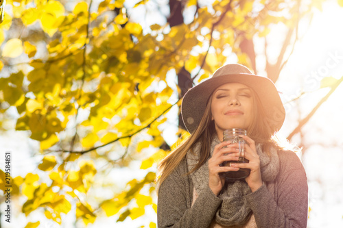 Young woman enjoying coffee outdoors on sunny autumn day. Closeup of female model standing by the tree with yellow leaves  with eyes closed holding coffee mug. Natural lighting  no retouch.