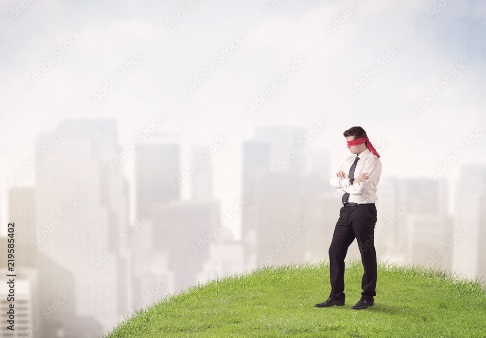 Young blindfolded businessman steps on a a patch of grass with a city in the background Young blindfolded businessman steps on a a patch of grass with a city in the background