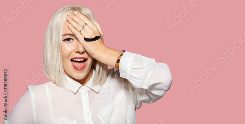 Young happy woman with eyelashes painted on the hand. Cute blonde hair model in white shirt closed her eye with hand. Concept of fun and merriment. Studio portrait on pink background.