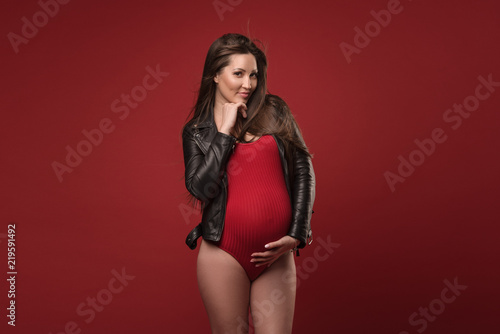 Fashionable pregnant woman with long healthy hair in stylish clothes on a red studio background. Carefully embraces his belly