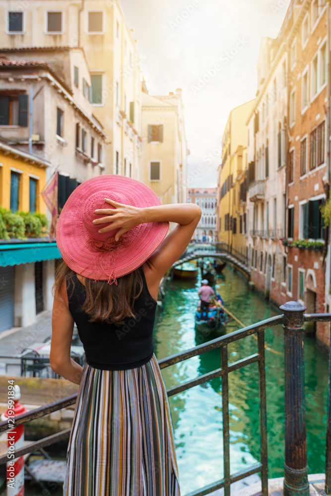 Fototapeta premium Elegancka kobieta z czerwonymi słońce kapeluszu spojrzeniami na kanale z przelotną gondolą w Wenecja, Włochy
