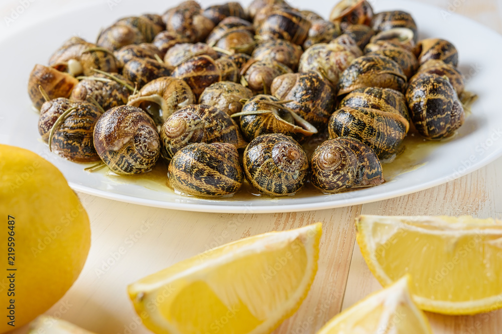 Tasty fried snails with lemon in white plate on white wooden background. Tasty fried snails with lemon in white plate on white wooden background.