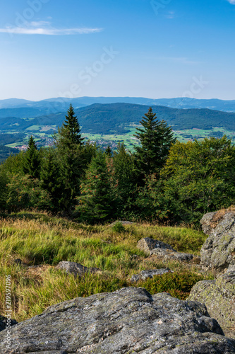 View from top of a mountain in the valley with clouds on the sky and mountains on the background and stones and trees in front of in the bavarian forest © Christian