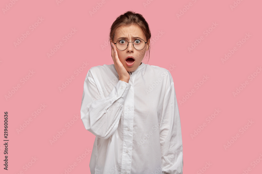 Stupefied displeased individual keeps mouth widely opened, touches cheeks with hand, dressed in white oversized shirt, poses against pink background. Omg concept. Horrified woman stands indoor Stupefied displeased individual keeps mouth widely opened, touches cheeks with hand, dressed in white oversized shirt, poses against pink background. Omg concept. Horrified woman stands indoor