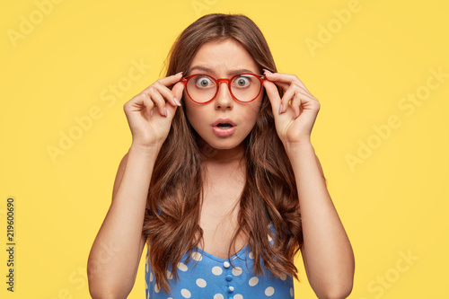 Fotografie Shocked attractive teenager wears glasses with red rim, polka dot blouse, stares at camera with frightened expression as hears horrified news, poses against yellow background