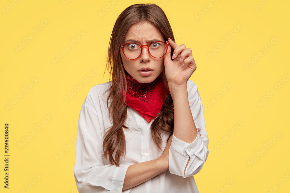 Oh no! Astounded young stylish woman stares with puzzlement and indignantion at camera, touches rim of spectacles, dressed in white oversized shirt and red bandana, doesnt believe in shocking news