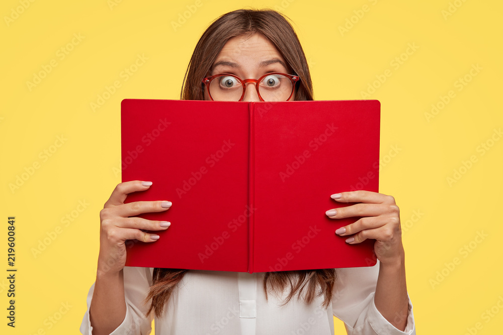 Stunned emotive student nerd hides face behind opened red book, wears glasses, does some tasks and prepares for classes in university, stands against yellow background. Girl enjoys reading literature Stunned emotive student nerd hides face behind opened red book, wears glasses, does some tasks and prepares for classes in university, stands against yellow background. Girl enjoys reading literature