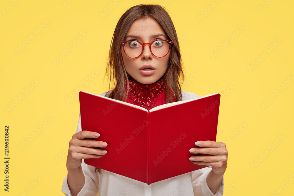 Youth, intelligence, knowledge, studying and emotions concept. Scared lovely schoolgirl holds red book, afraids of tomorrows exam at college, has fearful expression, isolated over yellow background. Youth, intelligence, knowledge, studying and emotions concept. Scared lovely schoolgirl holds red book, afraids of tomorrows exam at college, has fearful expression, isolated over yellow background.