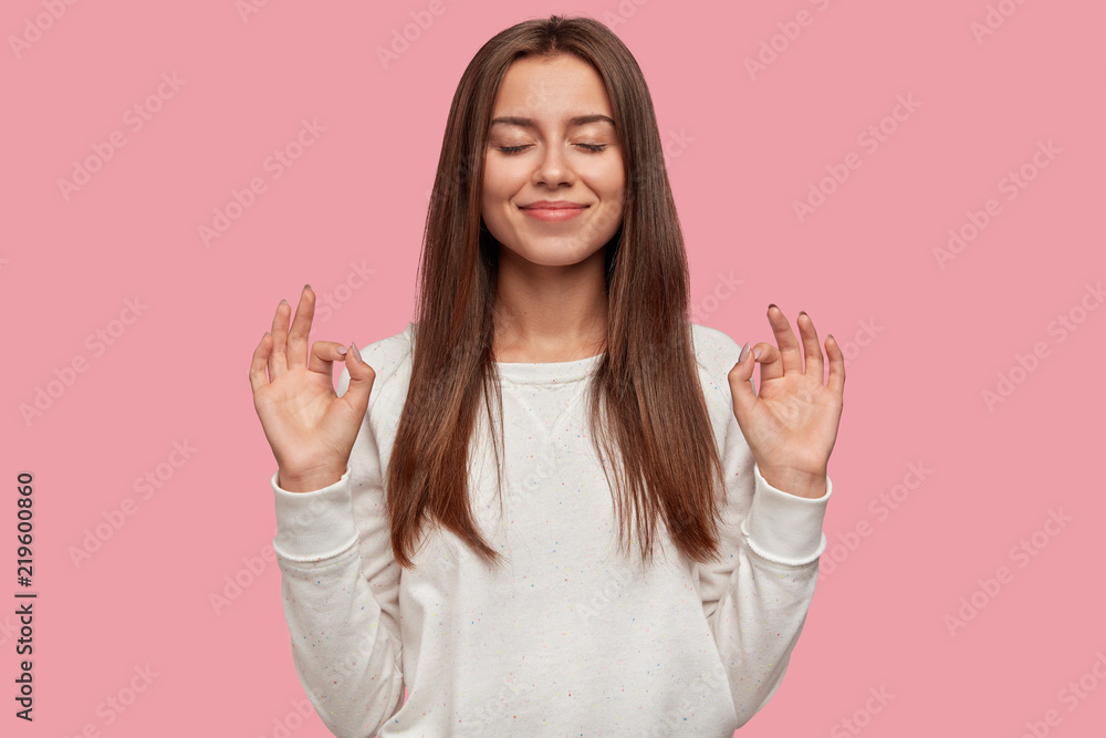 Pleased cheerful brunette woman makes okay gesture with both hands, says that everything is fine, closes eyes, dressed in white sweater, isolated over pink background. Body language and people concept Pleased cheerful brunette woman makes okay gesture with both hands, says that everything is fine, closes eyes, dressed in white sweater, isolated over pink background. Body language and people concept