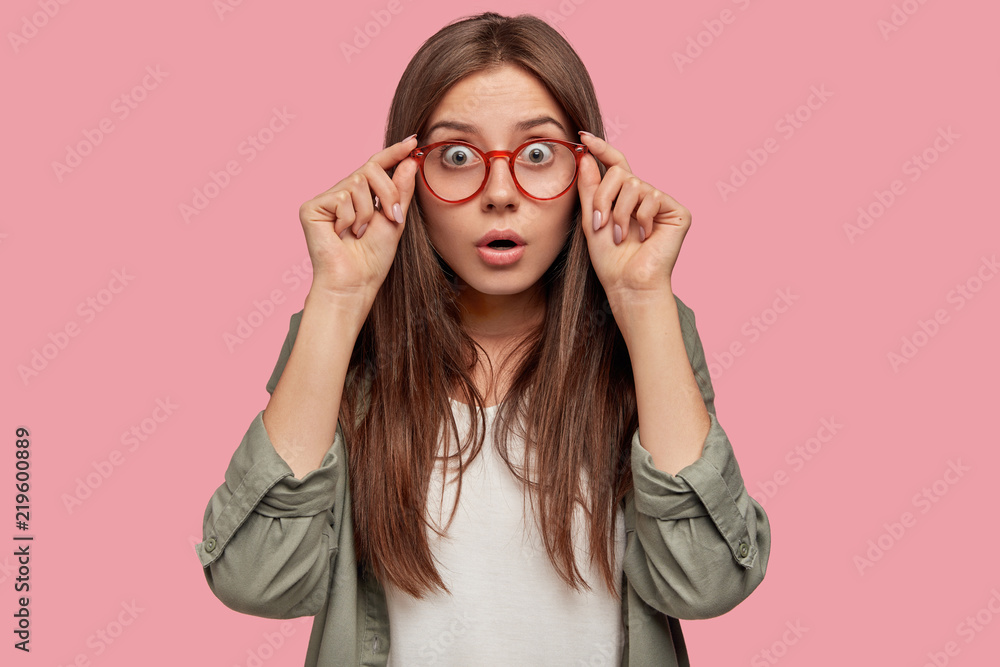Studio shot of astonished shocked woman stares through spectacles, wears casual shirt, listens her colleague at office, recieves unexpected news, stands against pink background. Reaction concept Studio shot of astonished shocked woman stares through spectacles, wears casual shirt, listens her colleague at office, recieves unexpected news, stands against pink background. Reaction concept
