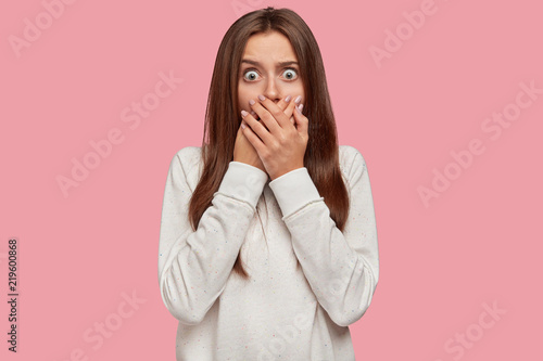 Frightened European woman covers mouth with both hands, recieves bad awful news, has dark straight hair, dressed in fashionable white sweater, isolated over pink background. People and astonishment © wayhome.studio 