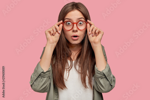 Quadro su tela Studio shot of astonished shocked woman stares through spectacles, wears casual shirt, listens her colleague at office, recieves unexpected news, stands against pink background