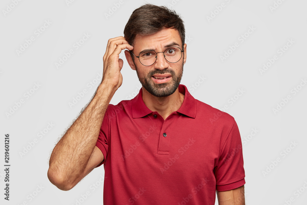 Puzzled unshaven guy scratches head, thinks deeply about something, has indignant look, dressed in casual clothes, stands against white background. Human facial expressions, emotions concept Puzzled unshaven guy scratches head, thinks deeply about something, has indignant look, dressed in casual clothes, stands against white background. Human facial expressions, emotions concept