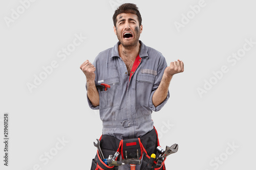 Photo of downcast stressed engineer clenches fists desperately, being despondent by bad work conditions, feels tired of constant hard labour, dressed in casual clothes, poses against white background