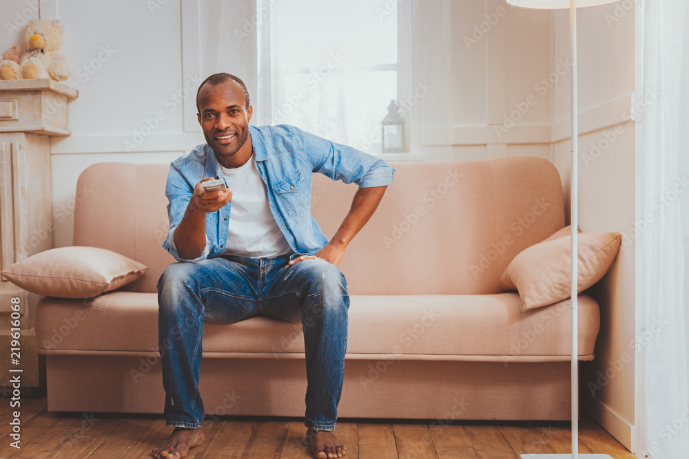 My day off. Content afro-american man sitting on the sofa and holding a  remote control Stock Photo | Adobe Stock