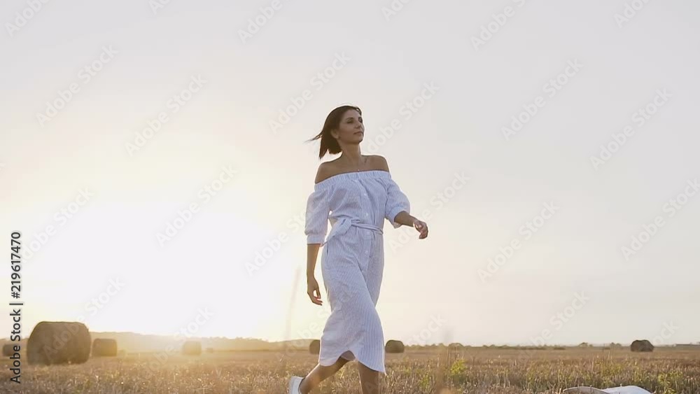 A young girl running in the field makes circles around him on a summer evening at sunset. Attractive girl in a long dress goes a trail on the field with bales of straw A young girl running in the field makes circles around him on a summer evening at sunset. Attractive girl in a long dress goes a trail on the field with bales of straw
